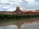 Glassy water, a band of dark green vegetation, with red cliffs and a rock tower in the distance